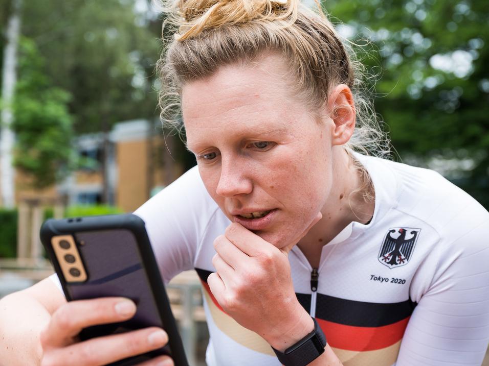 Athletin im weißen Trikot mit schwarzem und rotem Streifen schaut konzentriert auf ihr Smartphone in einer Außenanlage.