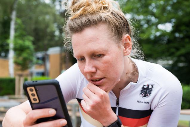 Athletin im weißen Trikot mit schwarzem und rotem Streifen schaut konzentriert auf ihr Smartphone in einer Außenanlage.