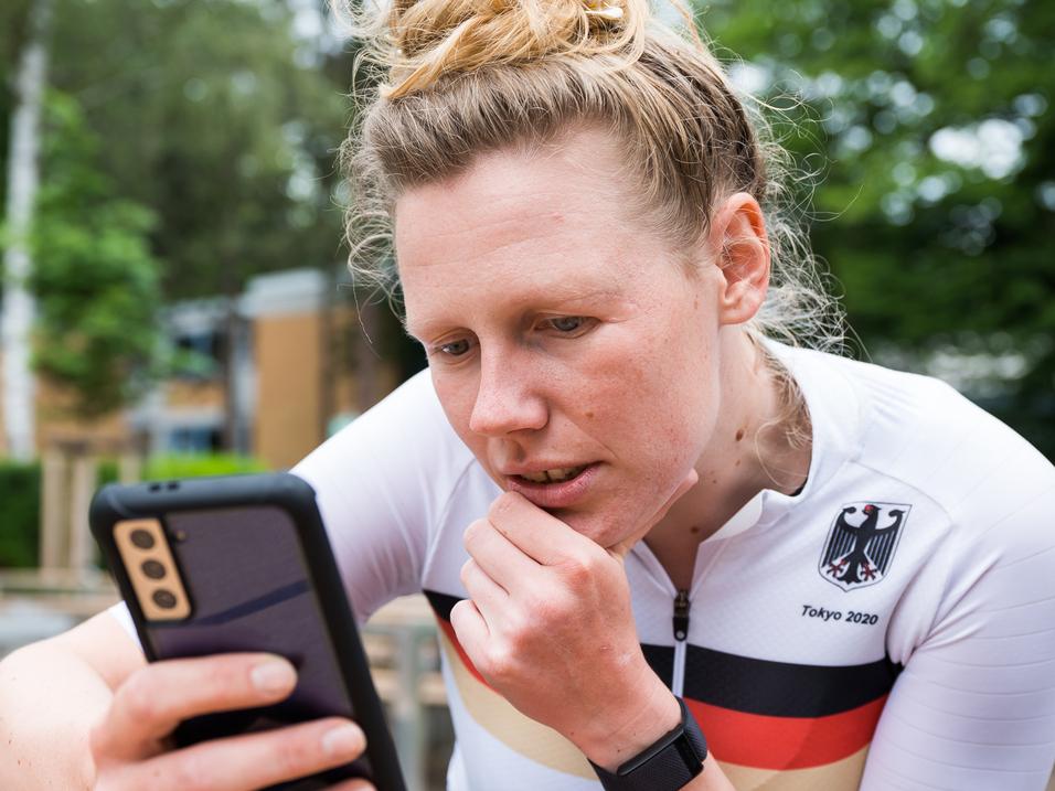 Athletin im weißen Trikot mit schwarzem und rotem Streifen schaut konzentriert auf ihr Smartphone in einer Außenanlage.